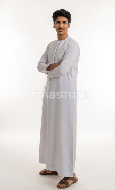 الاهتمام بالمظهر الخارجي, مفهوم الأناقة و الرجولة, تعابير الثقة بالنفس, بورتريه لشاب مراهق عربي خليجي إماراتي يرتدي الكندورة يقف باستقامة وأيدي متقاطعة وينظر إلى الكاميرا بإيماءات الاعتزاز بالذات, الجسم بالطول الكامل, خلفية بيضاء