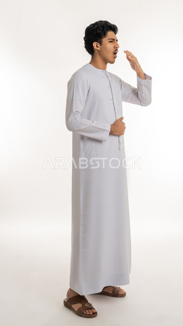 Need for sleep and rest, feeling tired and exhausted, expressions of laziness and lethargy, staying up late, portrait of a young Emirati Gulf Arab teenager wearing a kandura, putting his hand on his mouth with gestures of yawning and drowsiness, full body, white background