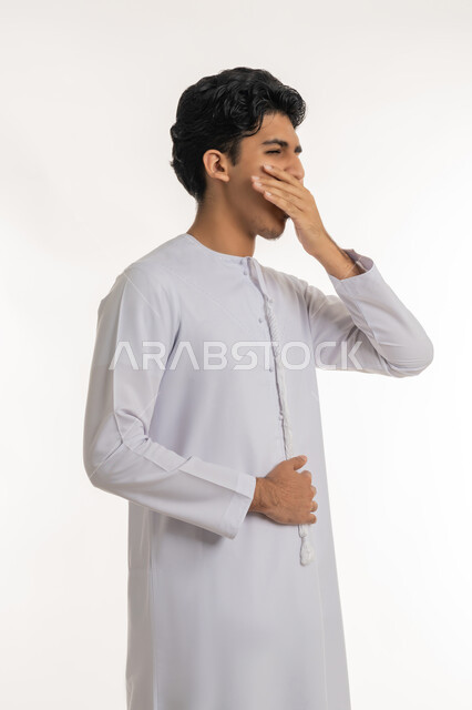 الإحساس بالإرهاق والتعب, السهر لوقت متأخر، تعابير الكسل والخمول، الحاجة للنوم والراحة, بورتريه لشاب مراهق عربي خليجي إماراتي يرتدي الكندورة يضع يده على فمه بإيماءات التثاؤب و النعاس, خلفية بيضاء