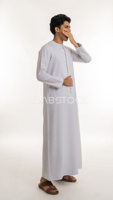 Need for sleep and rest, feeling tired and exhausted, expressions of laziness and lethargy, staying up late, portrait of a young Emirati Gulf Arab teenager wearing a kandura, putting his hand on his mouth with gestures of yawning and drowsiness, full body, white background
