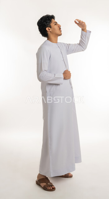 عسر الهضم والشعور بالمغص, الإحساس بآلام في المعدة, طلب المساعدة للذهاب إلى الطبيب، النظر إلى شيء ما في الأعلى، بورتريه لشاب مراهق عربي خليجي إماراتي يرتدي الكندورة يضع يده على بطنه بإيماءات الألم, الجسم بالطول الكامل, خلفية بيضاء