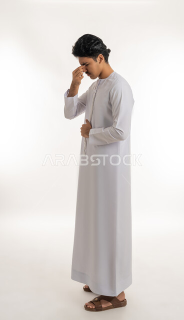 الصداع و آلام الرأس, الحاجة للفحص والعلاج، التهاب القرنية العينية, مشاكل في النظر, بورتريه لشاب مراهق عربي خليجي إماراتي يرتدي الكندورة يضع يده على وجهه بتعابير الألم, الجسم بالطول الكامل, خلفية بيضاء