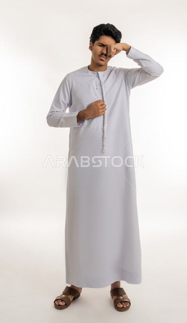 Expressions of laziness and lethargy, need for sleep and rest, feeling tired and exhausted, staying up late, portrait of a young Arab Gulf Emirati teenager wearing a kandura, putting his hand over his eyes with gestures of sleepiness, full body, white background