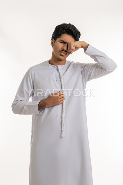 السهر لوقت متأخر، تعابير الكسل والخمول، الحاجة للنوم والراحة, الإحساس بالإرهاق والتعب, بورتريه لشاب مراهق عربي خليجي إماراتي يرتدي الكندورة يضع يده على عينه بإيماءات النعاس, خلفية بيضاء
