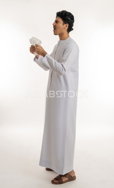 Allergic rhinitis and sinusitis, need for treatment and medication, feeling sick and cold, portrait of a young Arab Gulf Emirati teenager wearing a kandura putting a tissue on his face with sneezing gestures, full body, white background
