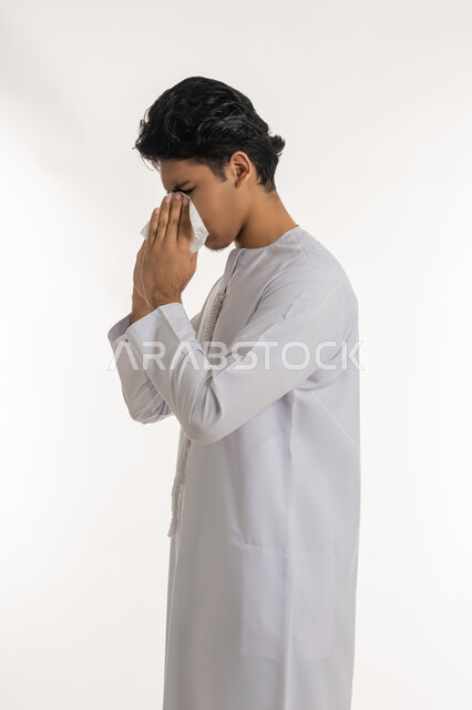 الشعور بالمرض و الزكام, الحاجة إلى العلاج والدواء, التهاب الأنف و الجيوب التحسسية, بورتريه لشاب مراهق عربي خليجي إماراتي يرتدي الكندورة يضع منديل ورقي على وجهه بإيماءات العطاس, خلفية بيضاء