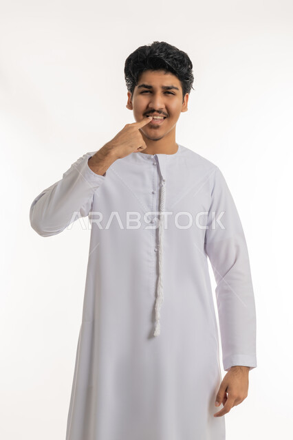 مفهوم تبييض وتجميل ومعالجة الأسنان, الاهتمام والعناية بنظافة وصحة الفم, تجنب التسوس وأمراض اللثة، بورتريه لشاب مراهق عربي خليجي إماراتي يرتدي الكندورة يشير بإصبع السبابة إلى أسنانه, خلفية بيضاء