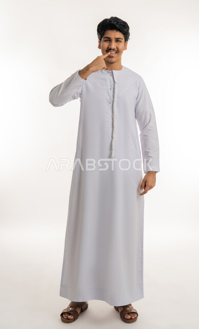 تجنب التسوس وأمراض اللثة, مفهوم تبييض وتجميل ومعالجة الأسنان, الاهتمام والعناية بنظافة وصحة الفم، بورتريه لشاب مراهق عربي خليجي إماراتي يرتدي الكندورة يشير بإصبع السبابة إلى أسنانه, الجسم بالطول الكامل, خلفية بيضاء