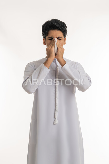 Feeling sick and cold, need for treatment and medicine, allergic rhinitis and sinusitis, portrait of a young Arab Gulf Emirati teenager wearing a kandura putting a tissue on his face with sneezing gestures, white background