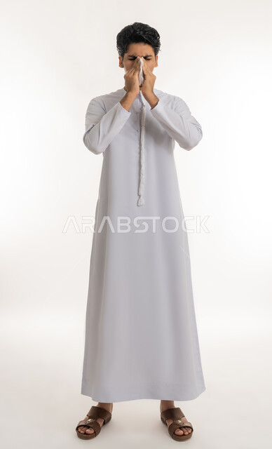 الحاجة إلى العلاج والدواء, التهاب الأنف و الجيوب التحسسية, الشعور بالمرض و الزكام, بورتريه لشاب مراهق عربي خليجي إماراتي يرتدي الكندورة يضع منديل ورقي على وجهه بإيماءات العطاس, الجسم بالطول الكامل, خلفية بيضاء