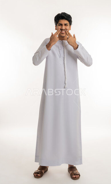 مفهوم معالجة و تجميل وتبييض الأسنان, الاهتمام والعناية بنظافة وصحة الفم, تجنب التسوس و أمراض اللثة، بورتريه لشاب مراهق عربي خليجي إماراتي يرتدي الكندورة يشير بأصابعه إلى أسنانه, الجسم بالطول الكامل, خلفية بيضاء