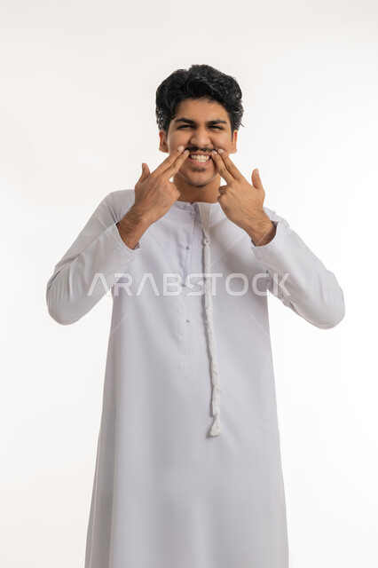 مفهوم معالجة و تجميل وتبييض الأسنان, الاهتمام والعناية بنظافة وصحة الفم, تجنب التسوس وأمراض اللثة، بورتريه لشاب مراهق عربي خليجي إماراتي يرتدي الكندورة يشير بأصابعه إلى أسنانه, خلفية بيضاء