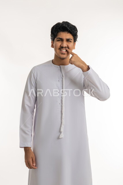 مفهوم تبييض وتجميل ومعالجة الأسنان, الاهتمام والعناية بنظافة وصحة الفم, تجنب التسوس وأمراض اللثة، بورتريه لشاب مراهق عربي خليجي إماراتي يرتدي الكندورة يشير بإصبع السبابة إلى أسنانه, خلفية بيضاء