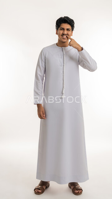 تجنب التسوس وأمراض اللثة, مفهوم تبييض وتجميل ومعالجة الأسنان, الاهتمام والعناية بنظافة وصحة الفم، بورتريه لشاب مراهق عربي خليجي إماراتي يرتدي الكندورة يشير بإصبع السبابة إلى أسنانه, الجسم بالطول الكامل, خلفية بيضاء