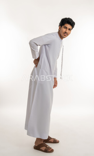 التهاب العضلات والمفاصل, آلام العامود الفقري والظهر, الأوجاع العصبية والعضلية، طلب المساعدة والحاجة للعلاج، بورتريه لشاب مراهق عربي خليجي إماراتي يرتدي الكندورة ينظر إلى الكاميرا و يضع يده على ظهره بإيماءات الألم, الجسم بالطول الكامل, خلفية بيضاء