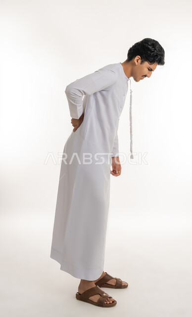 الأوجاع العصبية والعضلية، طلب المساعدة والحاجة للعلاج، التهاب العضلات والمفاصل, آلام العامود الفقري والظهر، بورتريه لشاب مراهق عربي خليجي إماراتي يرتدي الكندورة يضع يده على ظهره بإيماءات الألم, الجسم بالطول الكامل, خلفية بيضاء