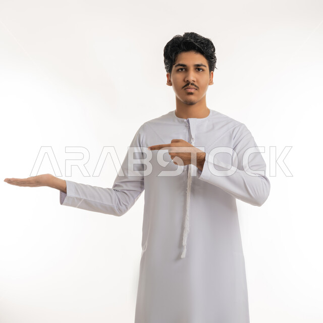 تعابير حمل و إمساك عنصر محدد, رفع شيء معين, الإشارة إلى هدف ما، بورتريه لشاب مراهق عربي خليجي إماراتي يرتدي الكندورة ينظر إلى الكاميرا و يشير بإصبع السبابة إلى يده، خلفية بيضاء