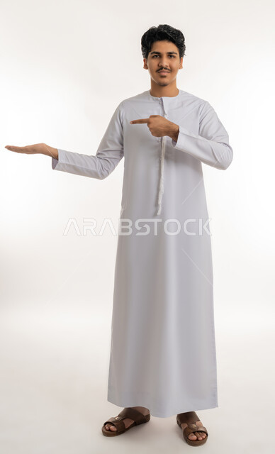 النظر إلى الكاميرا بإيماءات السعادة و السرور, رفع شيء معين, تعابير حمل و إمساك عنصر محدد, الإشارة إلى هدف ما، بورتريه لشاب مراهق عربي خليجي إماراتي يرتدي الكندورة يشير بإصبع السبابة إلى يده, الجسم بالطول الكامل، خلفية بيضاء