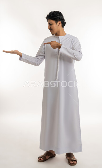 رفع شيء معين, تعابير حمل و إمساك عنصر محدد, الإشارة إلى هدف ما، بورتريه لشاب مراهق عربي خليجي إماراتي يرتدي الكندورة ينظر إلى يده و يشير بإصبع السبابة إليها, الجسم بالطول الكامل، خلفية بيضاء