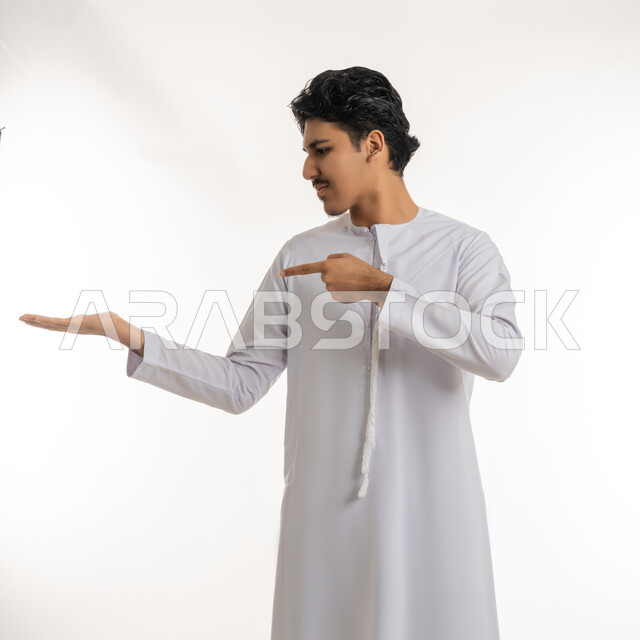رفع شيء معين, تعابير حمل و إمساك عنصر محدد, الإشارة إلى هدف ما، بورتريه لشاب مراهق عربي خليجي إماراتي يرتدي الكندورة ينظر إلى يده و يشير بإصبع السبابة إليها، خلفية بيضاء