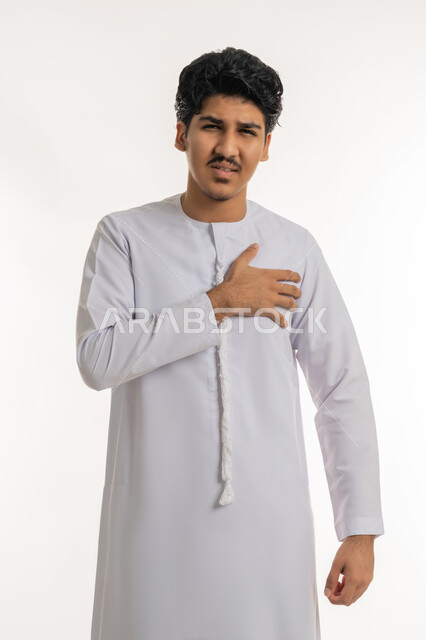 التعب والإرهاق والضغوطات النفسية، اضطرابات وأمراض قلبية, الشعور في زيادة معدل ضربات القلب, الإحساس بذبحة صدرية, بورتريه لشاب مراهق عربي خليجي إماراتي يرتدي الكندورة يضع يده على صدره بإيماءات الألم, خلفية بيضاء