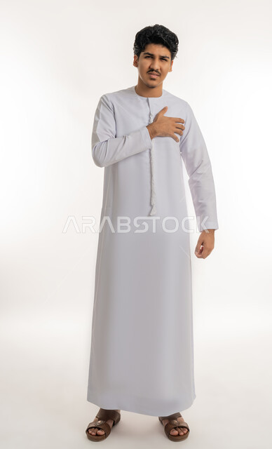 Heart disorders and diseases, feeling of increased heart rate, feeling of angina, fatigue, exhaustion and psychological pressures, portrait of a young Arab Gulf Emirati teenager wearing a kandura, putting his hand on his chest with gestures of pain, full-length body, white background