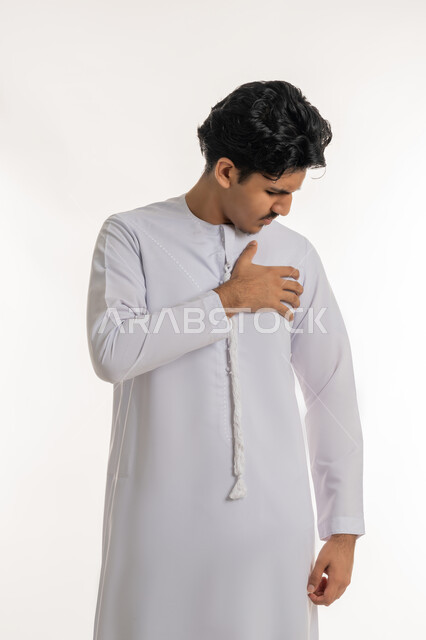 Feeling of angina, fatigue, exhaustion and psychological pressures, heart disorders and diseases, feeling of increased heart rate, portrait of a young Arab Gulf Emirati teenager wearing a kandura, putting his hand on his chest with gestures of pain, white background