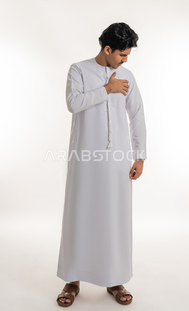Heart disorders and diseases, feeling of increased heart rate, feeling of angina, fatigue, exhaustion and psychological pressures, portrait of a young Arab Gulf Emirati teenager wearing a kandura, putting his hand on his chest with gestures of pain, full-length body, white background