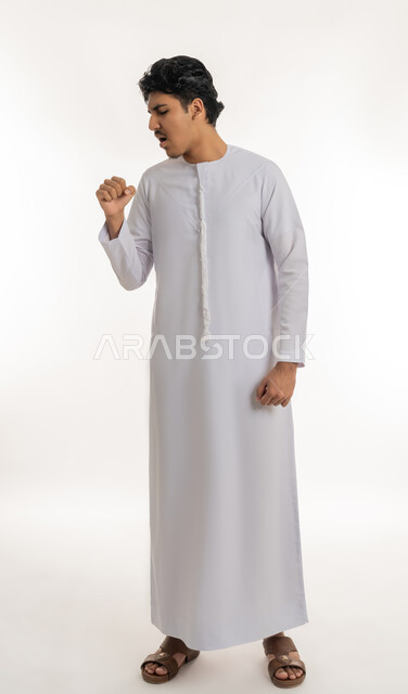 الشعور بالمرض و الكحة, الحاجة إلى زيارة الطبيب و العلاج, الإصابة بفايروس كورونا, الانفلونزا والتهاب الرئة, بورتريه لشاب مراهق عربي خليجي إماراتي يرتدي الكندورة يرفع يده إلى فمه بإيماءات السعال, الجسم بالطول الكامل, خلفية بيضاء