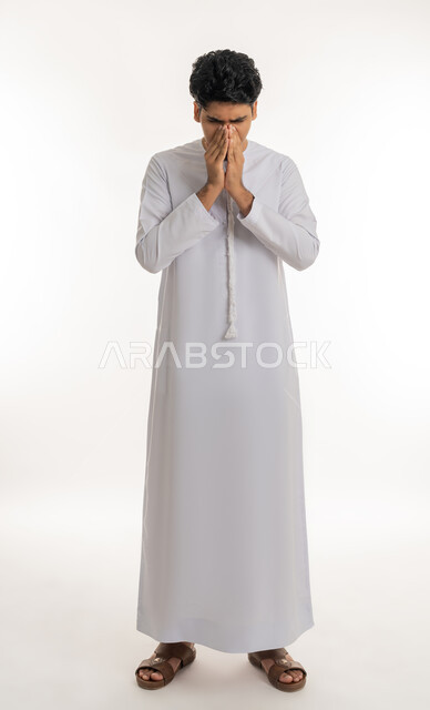 Feeling sick and cold, need for treatment and medicine, allergic rhinitis and sinusitis, portrait of a young Arab Gulf Emirati teenager wearing a kandura putting his hands on his face with sneezing gestures, full body, white background