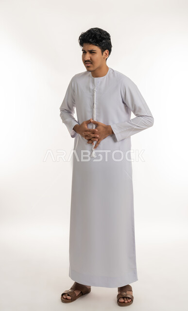 الحاجة للدواء ووضع خطة علاجية, عسر الهضم والشعور بالمغص, الإحساس بآلام في المعدة, طلب المساعدة للذهاب إلى الطبيب، بورتريه لشاب مراهق عربي خليجي إماراتي يرتدي الكندورة يضع يديه على بطنه بإيماءات الألم, الجسم بالطول الكامل, خلفية بيضاء