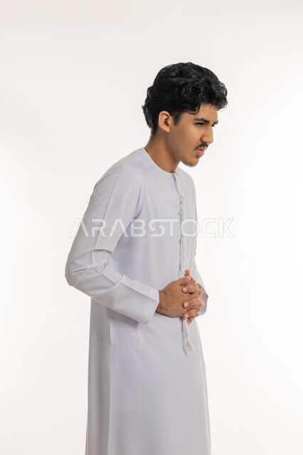 Indigestion and feeling of colic, feeling stomach pain, asking for help to go to the doctor, need for medication and a treatment plan, portrait of a young Arab Gulf Emirati teenager wearing a kandura putting his hands on his stomach with gestures of pain, white background