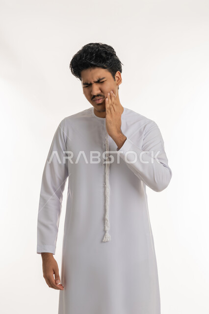 الحاجة لزيارة الطبيب, تسوس الأضراس وإلتهاب اللثة، طلب العلاج و المساعدة، بورتريه لشاب مراهق عربي خليجي إماراتي يرتدي الكندورة يضع يده على خده بإيماءات الشعور بألم الأسنان, خلفية بيضاء