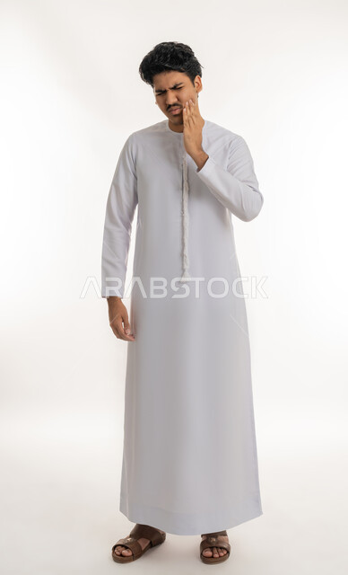 طلب العلاج و المساعدة, تسوس الأضراس وإلتهاب اللثة, الحاجة لزيارة الطبيب، بورتريه لشاب مراهق عربي خليجي إماراتي يرتدي الكندورة يضع يده على خده بإيماءات الشعور بألم الأسنان, الجسم بالطول الكامل, خلفية بيضاء