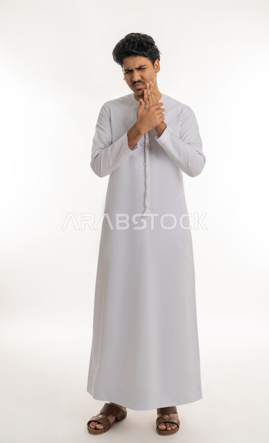 تسوس الأضراس وإلتهاب اللثة، طلب العلاج و المساعدة, الحاجة لزيارة الطبيب، بورتريه لشاب مراهق عربي خليجي إماراتي يرتدي الكندورة يضع يده على خده بإيماءات الشعور بألم الأسنان, الجسم بالطول الكامل, خلفية بيضاء