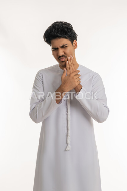 Need to visit a doctor, tooth decay and gum inflammation, seeking treatment and help, portrait of a young Emirati Gulf Arab teenager wearing a kandura, putting his hand on his cheek with gestures of feeling toothache, white background