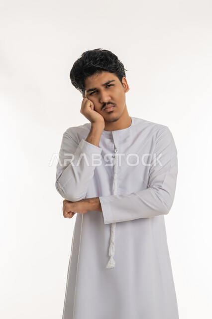 التفكير لإيجاد حل مشكلة ما, شرود الذهن والتأمل والتخطيط للمستقبل، مواجهة المشكلات والتحديات, بورتريه لشاب مراهق عربي خليجي إماراتي يرتدي الكندورة ينظر إلى الكاميرا و يضع يده على خده بتعابير التركيز و التمعن, خلفية بيضاء
