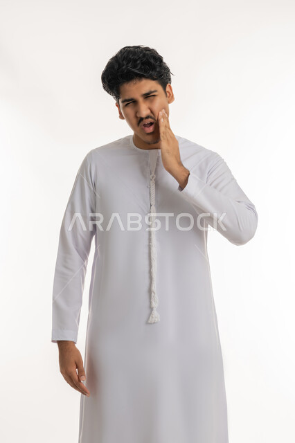 طلب العلاج و المساعدة, الحاجة لزيارة الطبيب, تسوس الأضراس وإلتهاب اللثة، بورتريه لشاب مراهق عربي خليجي إماراتي يرتدي الكندورة يضع يده على خده بإيماءات الشعور بألم الأسنان, خلفية بيضاء