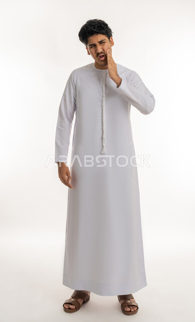 تسوس الأضراس وإلتهاب اللثة، طلب العلاج و المساعدة, الحاجة لزيارة الطبيب، بورتريه لشاب مراهق عربي خليجي إماراتي يرتدي الكندورة يضع يده على خده بإيماءات الشعور بألم الأسنان, الجسم بالطول الكامل, خلفية بيضاء