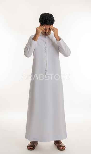 الحاجة إلى العلاج و الراحة, الشعور بالمرض و  التعب, آلام الشقيقة والصداع النصفي, الإحساس بالدوار و وجع الرأس, بورتريه لشاب مراهق عربي خليجي إماراتي يرتدي الكندورة يضع يديه على رأسه بإيماءات الانزعاج و الألم, الجسم بالطول الكامل, خلفية بيضاء
