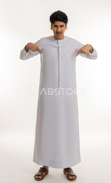 تأكيد حدوث شيء هذه اللحظة, حركات تدل على هذا المكان (هنا)، تحديد موقع معين، بورتريه لشاب مراهق عربي خليجي إماراتي يرتدي الكندورة ينظر إلى الكاميرا و يشير بإصابع السبابة نحو الأسفل, الجسم بالطول الكامل, خلفية بيضاء