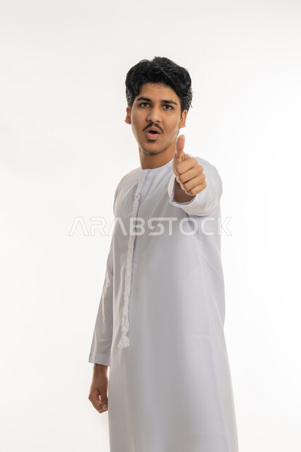 مفهوم القبول و الموافقة, تعابير الإعجاب و الرضى, بورتريه لشاب مراهق عربي خليجي إماراتي يرتدي الكندورة ينظر إلى الكاميرا و يرفع إصبع الإبهام للأعلى بإيماءات الجودة و الامتياز, خلفية بيضاء