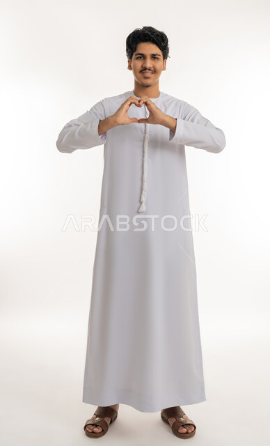 إظهار مشاعر الحب و الشغف، مفهوم المحبة و المودة, النظر إلى الكاميرا بإيماءات السعادة و السرور, بورتريه لشاب مراهق عربي خليجي إماراتي يرتدي الكندورة يشكل بيديه شكل قلب, الجسم بالطول الكامل, خلفية بيضاء