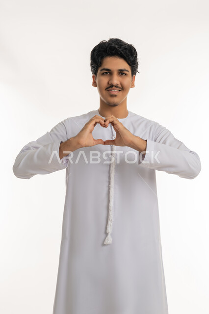 إظهار مشاعر الحب و الشغف، مفهوم المحبة و المودة, النظر إلى الكاميرا بإيماءات السعادة و السرور, بورتريه لشاب مراهق عربي خليجي إماراتي يرتدي الكندورة يشكل بيديه شكل قلب, خلفية بيضاء