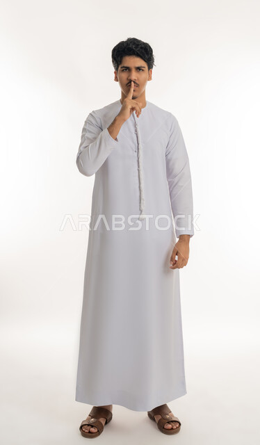 التوقف عن الكلام و الأمر بالصمت و عدم إصدار صوت, النظر إلى الكاميرا بإيماءات الجدية و الحزم، بورتريه لشاب مراهق عربي خليجي إماراتي يرتدي الكندورة يرفع إصبع السبابة إلى فمه بتعابير طلب الهدوء, الجسم بالطول الكامل, خلفية بيضاء