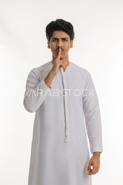 التوقف عن الكلام و الأمر بالصمت و عدم إصدار صوت, النظر إلى الكاميرا بإيماءات الجدية و الحزم، بورتريه لشاب مراهق عربي خليجي إماراتي يرتدي الكندورة يرفع إصبع السبابة إلى فمه بتعابير طلب الهدوء, خلفية بيضاء