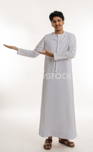 Gestures of lifting and carrying an item, greeting and welcoming and receiving a person, portrait of a young Arab Gulf Emirati teenager wearing a kandura looking at the camera with gestures of happiness and pleasure and pointing towards a place, full body, white background