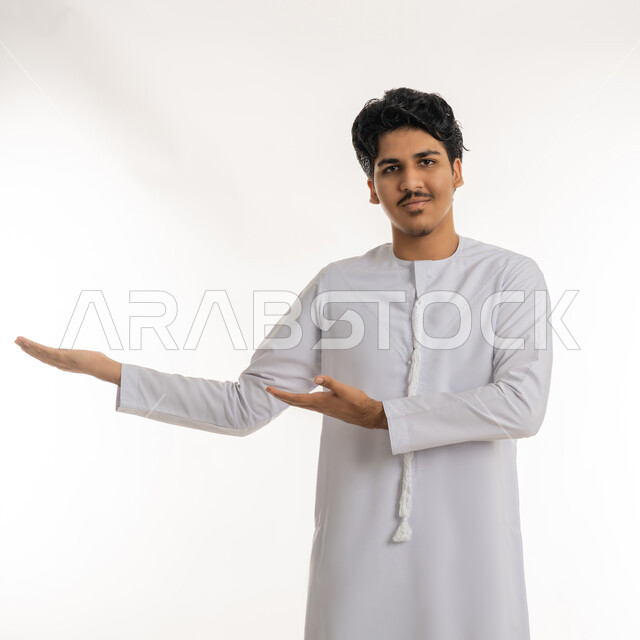 إشارات رفع و حمل عنصر ما, التحية والترحيب و استقبال شخص، بورتريه لشاب مراهق عربي خليجي إماراتي يرتدي الكندورة ينظر إلى الكاميرا بإيماءات السعادة و السرور و يشير نحو مكان ما، خلفية بيضاء