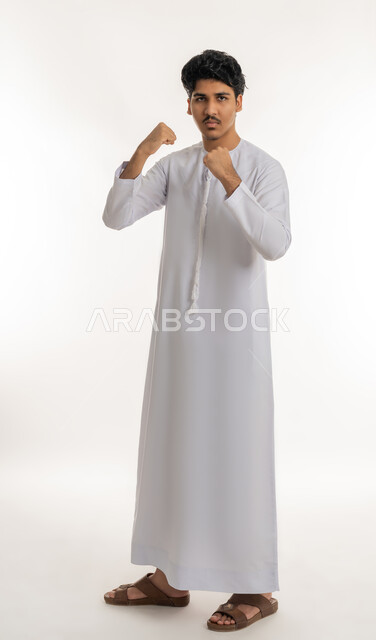 التدرب على حركات قتالية خاصة بالكارتيه، تعابير القوة و المنافسة والتحدي, ممارسة رياضة الدفاع عن النفس، بورتريه لشاب مراهق عربي خليجي إماراتي يرتدي الكندورة يرفع قبضة يديه للإستعداد للقتال, الجسم بالطول الكامل، خلفية بيضاء