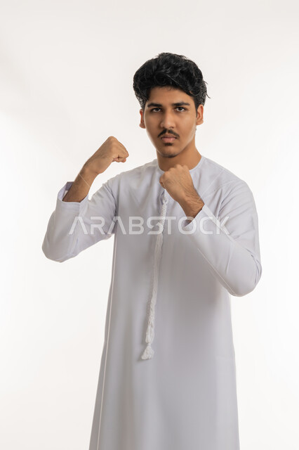 التدرب على حركات قتالية خاصة بالكارتيه، تعابير القوة و المنافسة والتحدي, ممارسة رياضة الدفاع عن النفس، بورتريه لشاب مراهق عربي خليجي إماراتي يرتدي الكندورة يرفع قبضة يديه للإستعداد للقتال، خلفية بيضاء